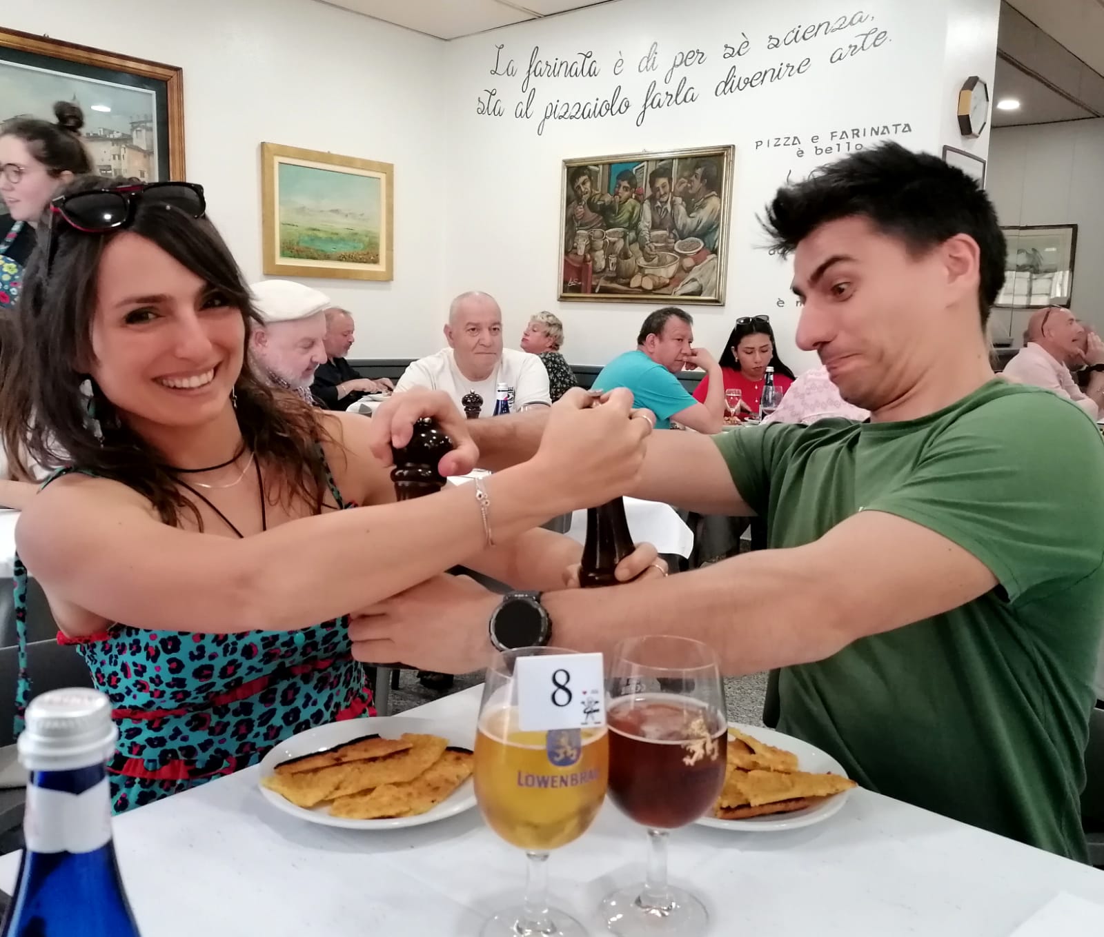 Two people at an Italian PIZZERIA, laughing while seasoning food with a pepper grinder, enjoying "FARINATA" traditional Italian food and beer